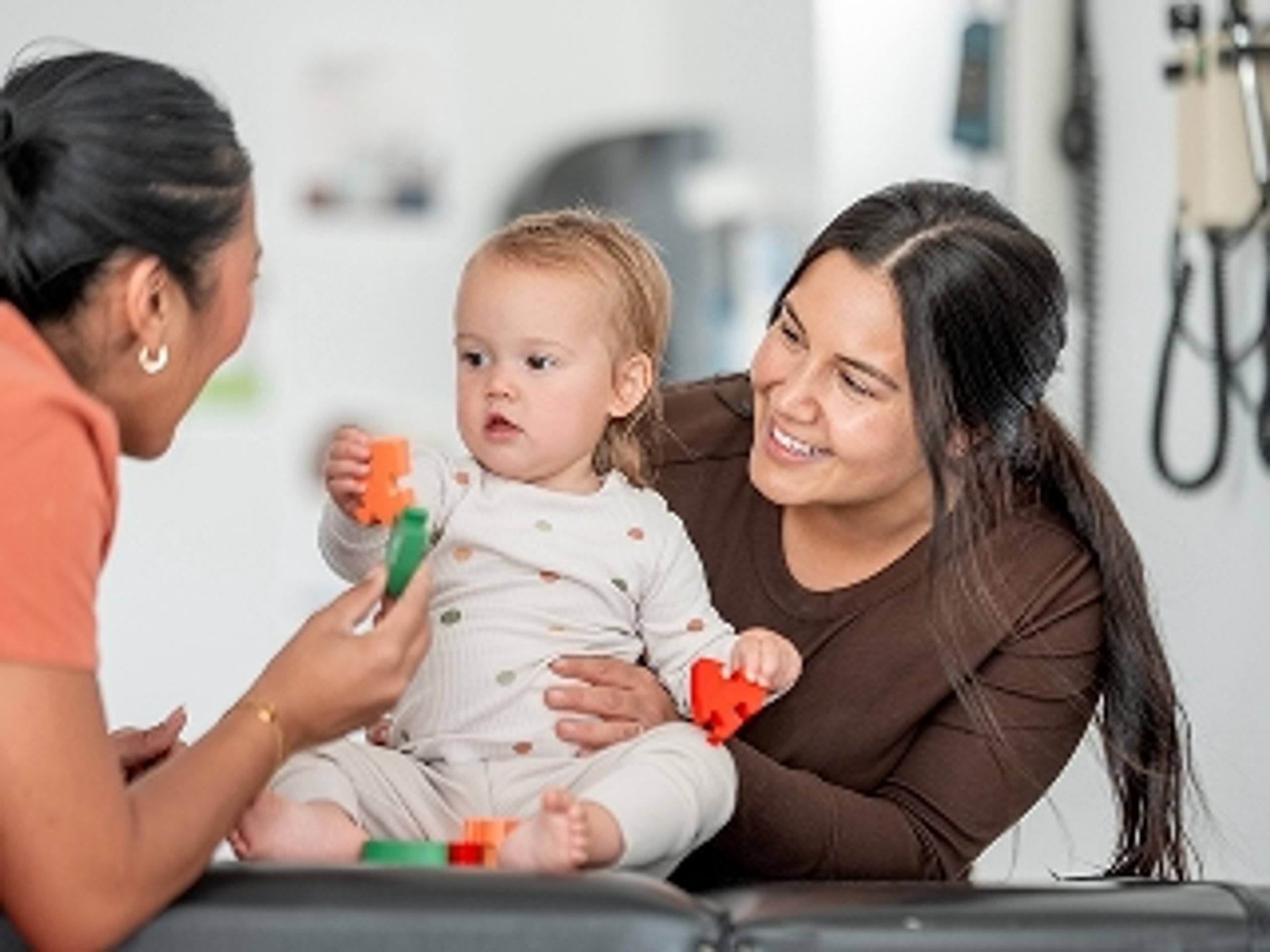 A woman is playing with a baby while another woman watches, both smiling. The baby is holding colourful toys, and they are in a bright, casual setting.